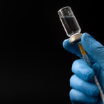 Insulin shot, flu jab or medical injection concept theme with doctor or nurse hands wearing blue surgical latex gloves filling a syringe from a glass vial isolated on black background with copyspace